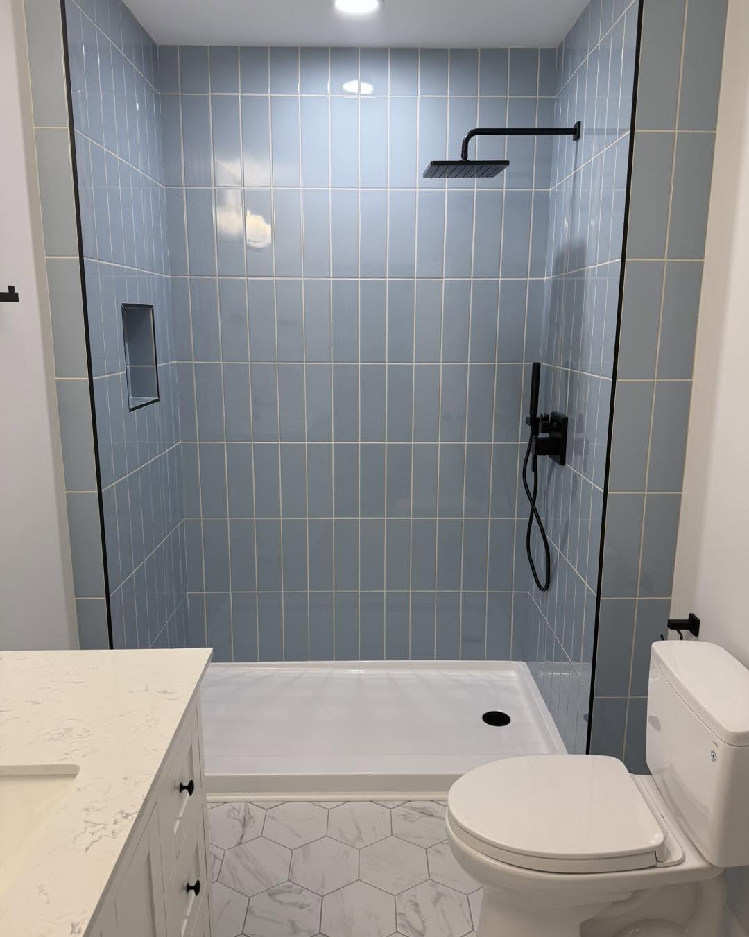 Blue tile walk-in shower with black fixtures and hexagon tile floor by Fisher Remodeling