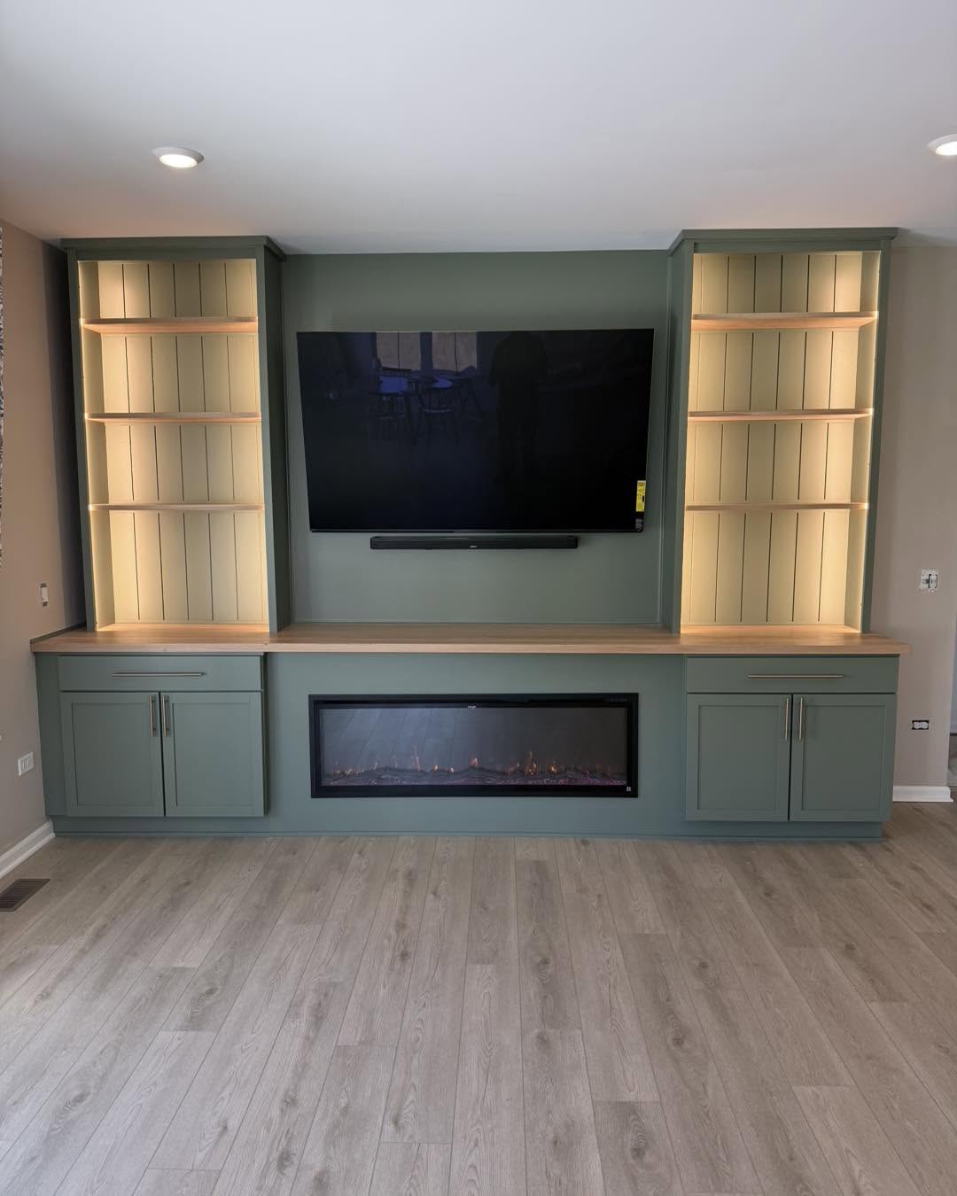 Sage green built-in entertainment wall with electric fireplace and lit shelving by Fisher Remodeling