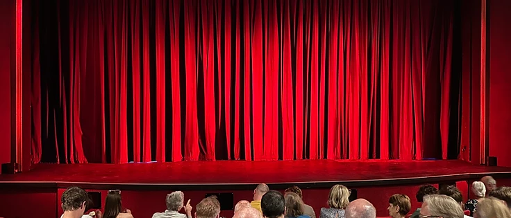Audience seated in a theatre auditorium facing a stage with red curtains.