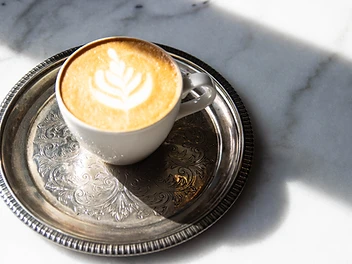 A latte with leaf-pattern latte art in a white cup on an ornate silver tray, photographed on marble at The Hive Store, Altrincham