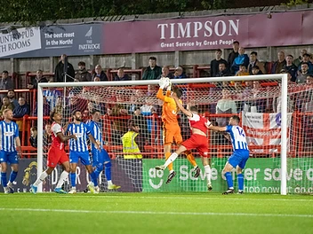 Goalmouth scramble at Altrincham FC under floodlights, with a goalkeeper in orange leaping to catch the ball and players challenging.