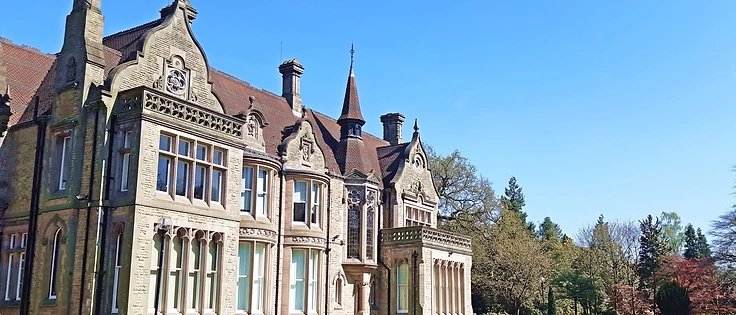 Denzell House, a Victorian-Gothic stone manor in Bowdon, with ornate gables, a central tower and manicured lawn under a clear blue sky.
