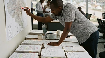 A campaign war room table in Nairobi is covered with annotated demographic research reports and a large wall map detailing local constituency boundaries.