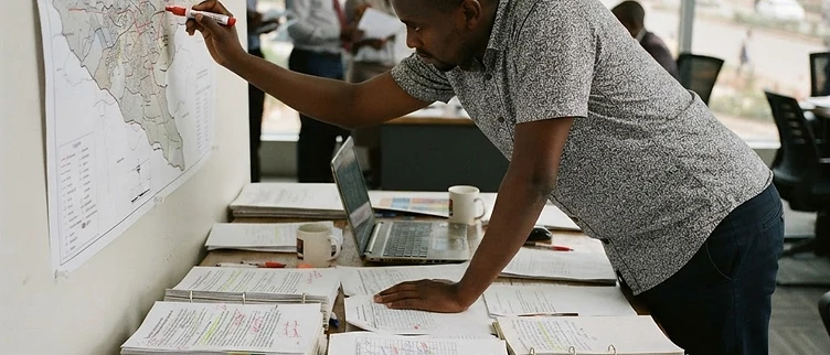 A campaign war room table in Nairobi is covered with annotated demographic research reports and a large wall map detailing local constituency boundaries.