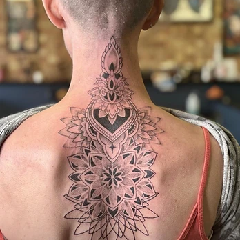 Fine-line black and grey ornamental mandala with dotwork shading and a spear-like peak, running down the nape of the neck and upper back