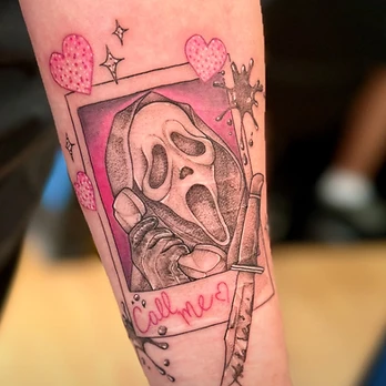 Fine-line black and grey tattoo of a masked screaming face holding a phone inside a Polaroid frame with a knife alongside, pink hearts and the words Call me?, on a forearm