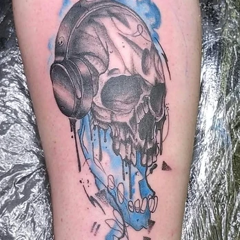 Black and grey tattoo of a skull in over-ear headphones, its jaw dissolving into a splash of blue ink with a thin line trailing to a small skull icon