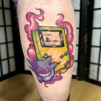 Colour tattoo of a lime green handheld games console with a pixel-art screen and text box, in front of a purple grinning ghost character wrapped in magenta smoke, on a lower leg
