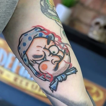 Japanese-inspired colour tattoo of a hyottoko-style mask with a winking eye, a spotted blue head wrap and red accents, on an upper arm