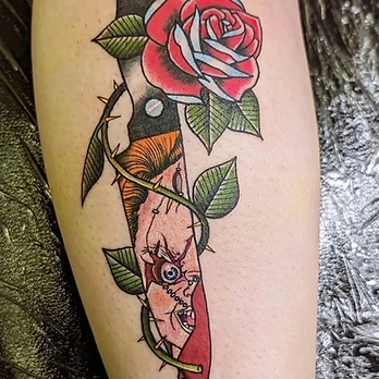 Colour horror tattoo of a kitchen knife blade carrying Chucky's stitched face, wrapped by a thorny vine and a bright red rose with light blue highlights