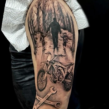 Black and grey tattoo on an upper arm showing a parent and two children walking through woodland above a motocross bike and crossed spanners