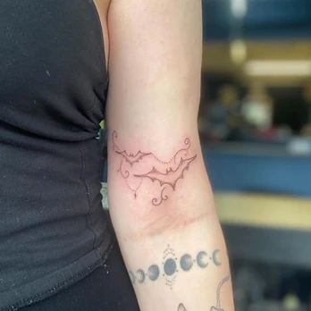 Fine line tattoo of two bats in flight joined by delicate lines and filigree flourishes, with a fine dotted chain and a tiny cross charm hanging below them, on an upper inner arm