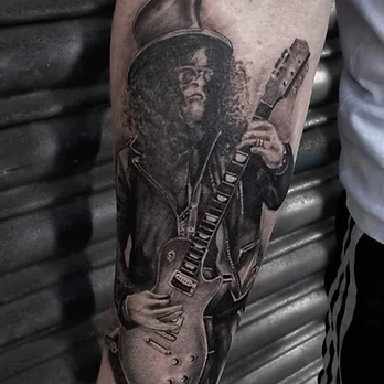 Black and grey portrait tattoo of the guitarist Slash in his top hat, curly hair and sunglasses playing an electric guitar, on a forearm