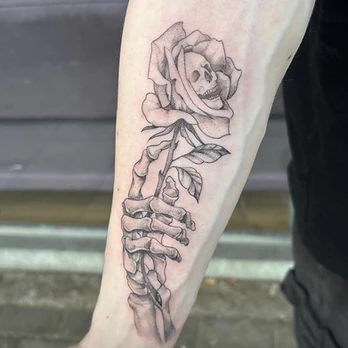 Fine-line black and grey tattoo of a skeleton hand reaching up to hold a long-stemmed rose, with a skull formed inside the petals, on a forearm