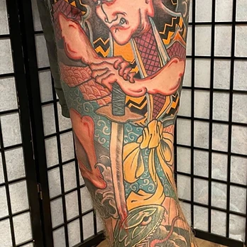 Large colour tattoo of a samurai holding a katana, framed by grey cloud above and a dark scaled snake winding through orange wave patterns, on an upper thigh