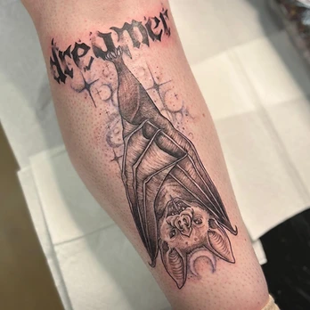 Fine-line black and grey tattoo of a bat hanging upside down above a crescent moon, with small stars and the word dreamer lettered above, on a forearm
