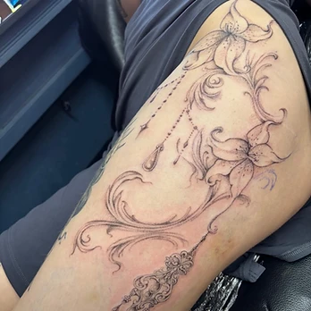 Fine-line black and grey tattoo of two botanical lilies joined by swirling filigree and a beaded chain ending in a teardrop pendant, above an ornate chandelier, on an upper thigh