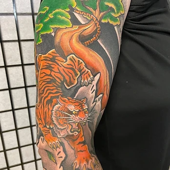 Traditional-style colour tattoo of an orange tiger climbing a tree with bright green foliage against dark shading, on an upper arm