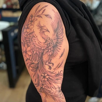 Fine-line black and grey tattoo of a phoenix with its wings raised and tail feathers falling, surrounded by five-petalled flowers, on an upper arm