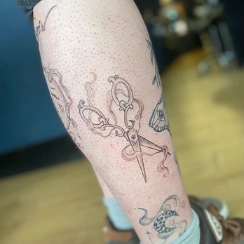 Fine line tattoo of a pair of ornate vintage tailoring scissors with filigreed handles, surrounded by curling wisps of smoke, on a lower leg