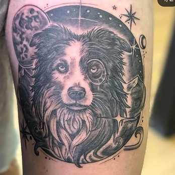 Black and grey tattoo of a border collie's face inside a circular frame, with a moon, stars and swirling cloud behind it, on an upper arm