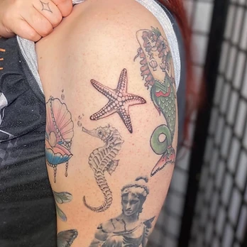 Fine line tattoo collage of a stippled starfish above a seahorse, with an open shell holding a coloured pearl beside a mermaid figure and a classical statue bust, on an upper arm