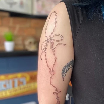 Fine line tattoo of a draped rosary bead chain with a heart-shaped centre link and a small crucifix, gathered with a ribbon tied in a bow, on an upper arm
