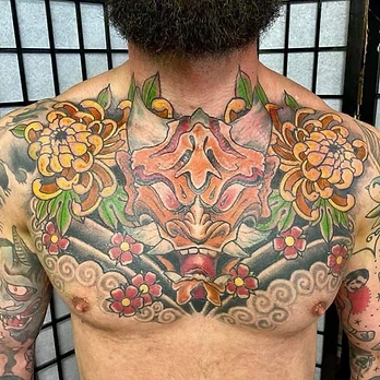 Japanese-style colour tattoo of a hannya mask in peach and orange, framed by yellow chrysanthemums and red blossom across a chest