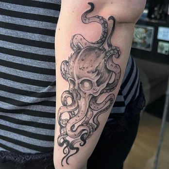 Fine-line black and grey tattoo of a cracked human skull with octopus tentacles twisting out from behind it, suction cups showing, on an upper arm