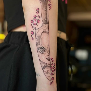 Fine-line black and grey tattoo of a scarred doll-like face set inside a chef's knife blade, flanked by pink leopard-print spots, on a lower forearm