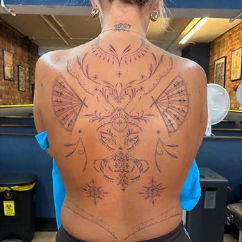 Fine-line black and grey ornamental back tattoo with matching fan motifs over each shoulder blade, a decorative column down the spine and geometric star mandalas at the base