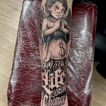 Black and grey tattoo of a winged cherub in street style with hands in prayer above a money bag, lettered One Life One Chance, on a forearm
