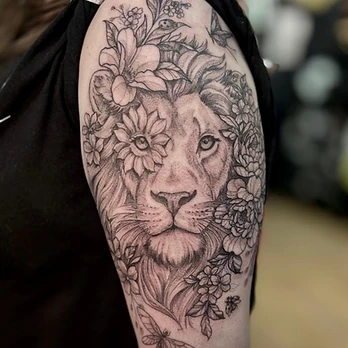 Fine-line black and grey tattoo of a lion's face with a large bloom over one eye, framed by blossoms and leaves with a dragonfly below, on an upper arm