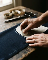 Primer plano de manos de artesano trazando una línea de patrón con tiza blanca sobre tela de lana azul marino, tijeras y cinta métrica visibles pero desenfocadas