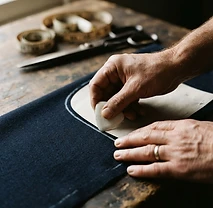 Primer plano de manos de artesano trazando una línea de patrón con tiza blanca sobre tela de lana azul marino, tijeras y cinta métrica visibles pero desenfocadas