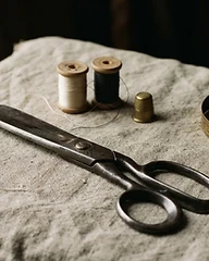 Naturaleza muerta de herramientas de atelier sobre lino prensado: tijeras de acero pesadas, carretes de hilo de seda, cinta métrica de bronce y una bobina de madera