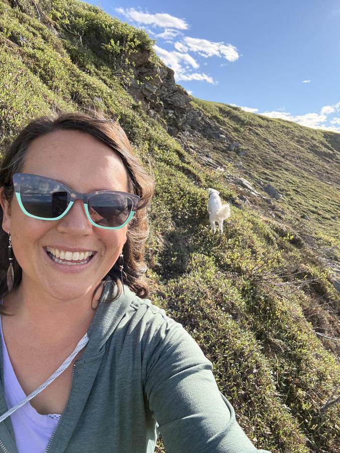 Dr. Alex smiling on an Alaska hillside with her white dog behind her