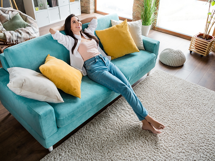 A woman leaning back relaxed on a teal couch in a bright, clutter-free living room, smiling at the ceiling