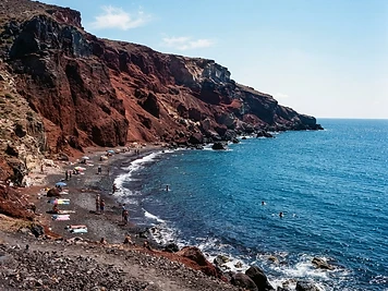 Rust-red volcanic cliffs above dark pebbles and blue water at Red Beach, Santorini.