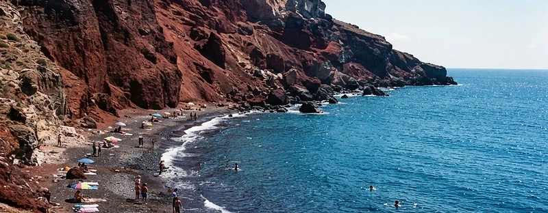 Rust-red volcanic cliffs above dark pebbles and blue water at Red Beach, Santorini.