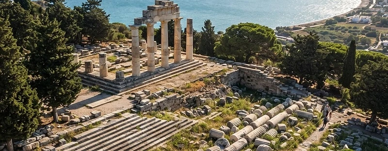 The terraces, steps and fallen columns of the Asklepion of Kos, with a wide bay below and cypresses on the slope.