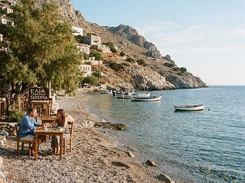 A narrow pebble shore with clear shallow water and taverna tables under a tamarisk at Vlichos, Hydra.