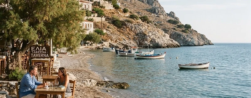 A narrow pebble shore with clear shallow water and taverna tables under a tamarisk at Vlichos, Hydra.
