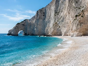The white pebble beach at Lalaria under white limestone cliffs on Skiathos, with a rock arch in the water.
