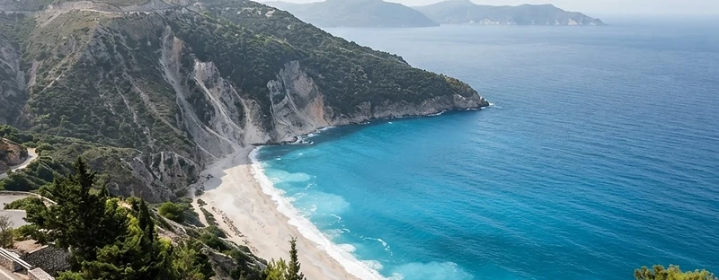 A crescent bay of turquoise water below white limestone cliffs on Kefalonia, seen from the coast road.
