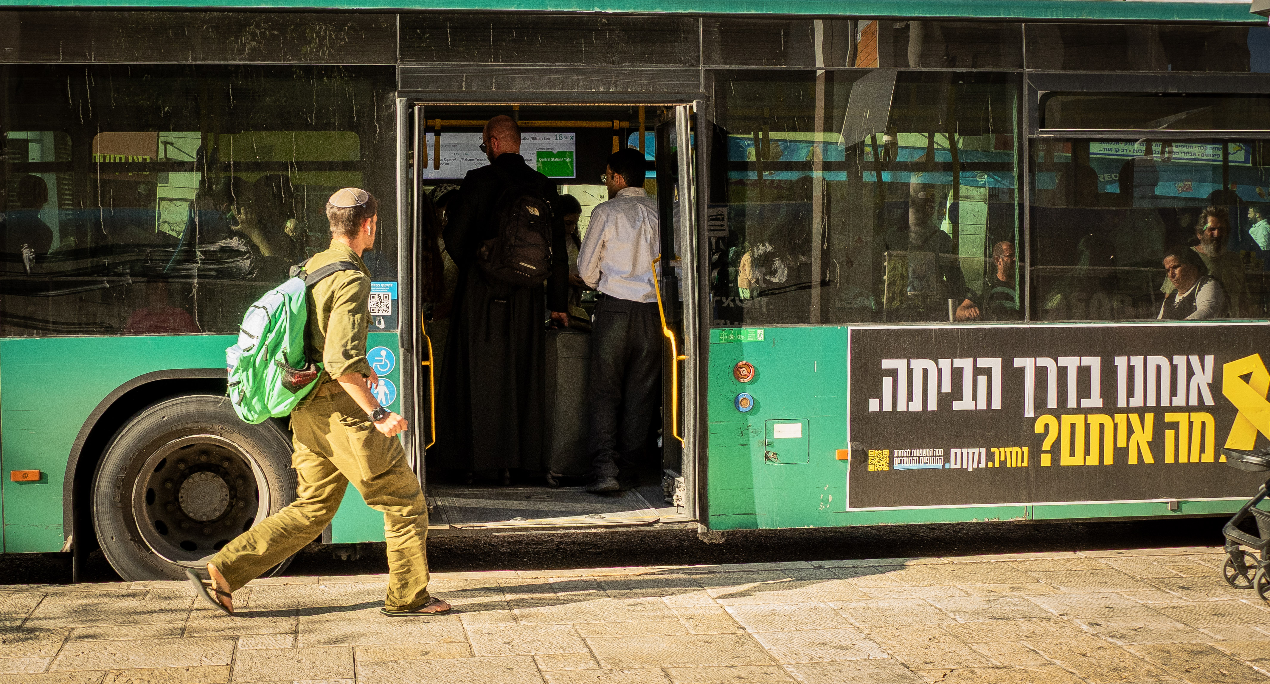 תיעוד רגע יומיומי בעולם, בו נראה חייל עם תיק גב בולט העולה לאוטובוס עירוני. התאורה הצידית מדגישה את הטקסטורות על דופן הרכב והדמויות, בעוד שהקומפוזיציה מנצלת את תנועת החייל ליצירת עניין. הפרטים הצילומיים משקפים רגע דוקומנטרי חי עם חדות טובה וצבעוניות מובהקת בתוך סביבה אורבנית אופיינית.