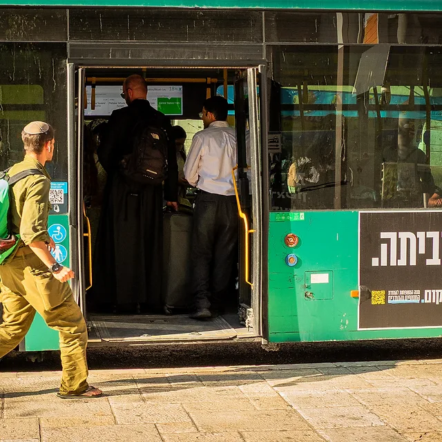 תיעוד רגע יומיומי בעולם, בו נראה חייל עם תיק גב בולט העולה לאוטובוס עירוני. התאורה הצידית מדגישה את הטקסטורות על דופן הרכב והדמויות, בעוד שהקומפוזיציה מנצלת את תנועת החייל ליצירת עניין. הפרטים הצילומיים משקפים רגע דוקומנטרי חי עם חדות טובה וצבעוניות מובהקת בתוך סביבה אורבנית אופיינית.