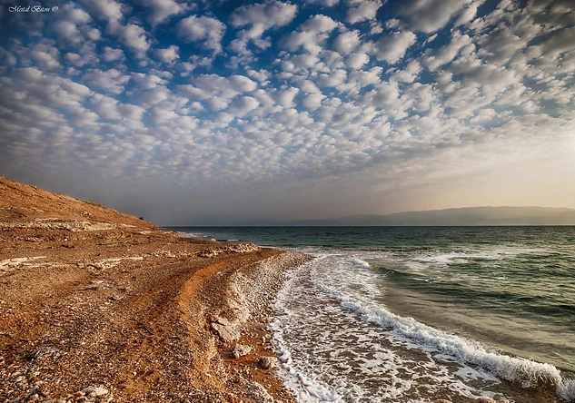 צילום נוף רחב המציג קו חוף מחוספס בעל טקסטורה של חלוקי נחל, הנמשך אל עבר אופק רחוק. השמיים מעוטרים בענני 'כסות' דרמטיים המעניקים עומק רב לקומפוזיציה. התאורה הרכה של שעת בין ערביים מדגישה את הניגודיות בין גווני האדמה החמים לבין המים בגווני כחול-ירוק, ויוצרת אווירה שלווה ומרגיעה.