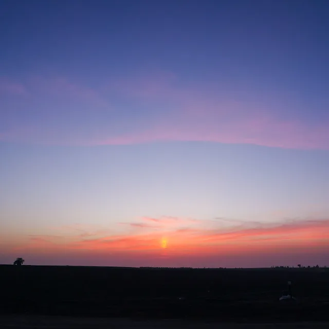צילום נוף מרהיב המציג שקיעה שקטה מעל אופק רחב. השמיים מתמזגים בגוונים הדרגתיים של כחול עמוק, ורוד עדין וכתום בוער סביב השמש השוקעת. הקומפוזיציה המינימליסטית מדגישה את המרחב הפתוח, כאשר קו הרקיע הכהה והדומם יוצר ניגודיות חזקה ומעניק לתמונה אווירה רגועה, פסטורלית ומלאה בהוד.