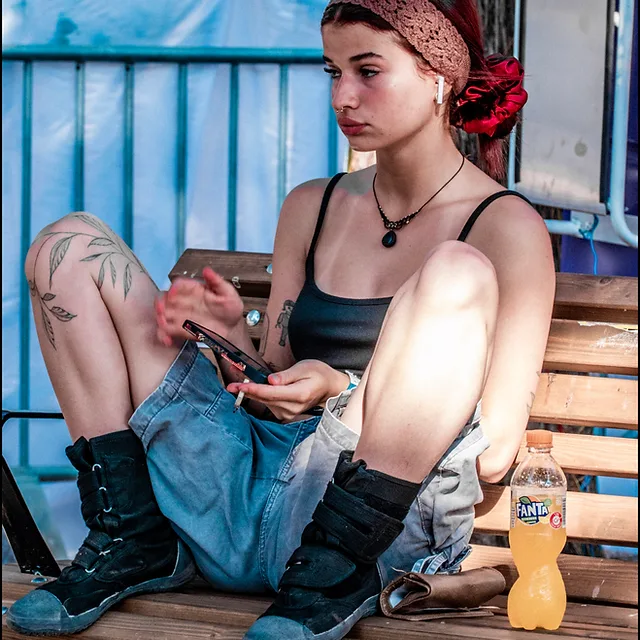 דיוקן דוקומנטרי צבעוני המציג צעירה יושבת על ספסל בעולם, כשהיא מחזיקה טלפון וסיגריה בידיה. הצילום שבוצע ב-Panasonic DMC-FZ1000 בתאורת יום טבעית, מתאפיין בחדות גבוהה על הדמות וביצירת קומפוזיציה המדגישה את המראה האורבני והקז'ואלי שלה, עם משחקי צבע של כחול, כתום ומרקמי עור וטקסטיל ברורים.